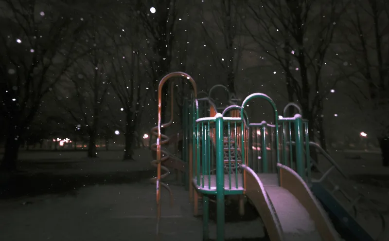 A snow-dusted playground climbing frame and slide in an unlit park at night, flakes falling through bare trees