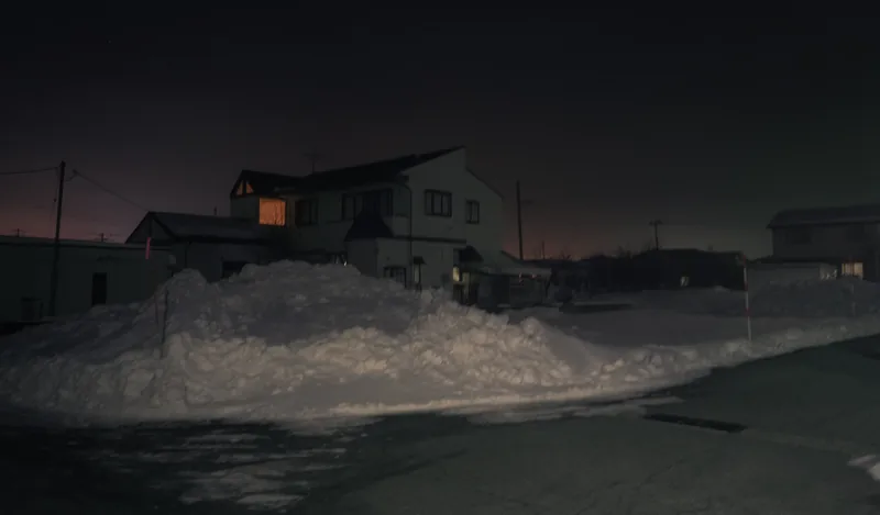 A cleared snowbank piled along an empty road in front of suburban houses, one warm second-storey window lit