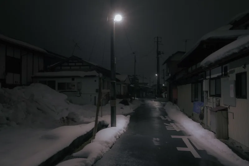 A narrow snow-lined Japanese street framed by houses, lit from above by a single streetlamp