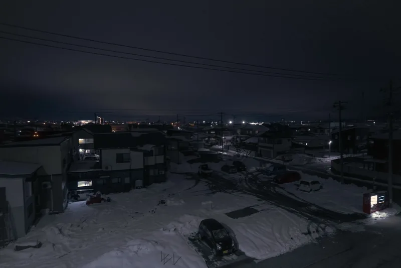 An elevated night view over a snow-covered parking lot and rooftops laced with power lines, a red vending machine glowing