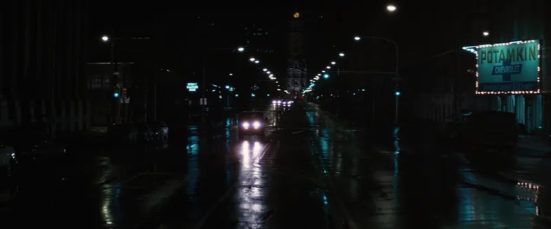 A single car on a dark wet boulevard drives toward an illuminated clock tower under glowing street lamps.