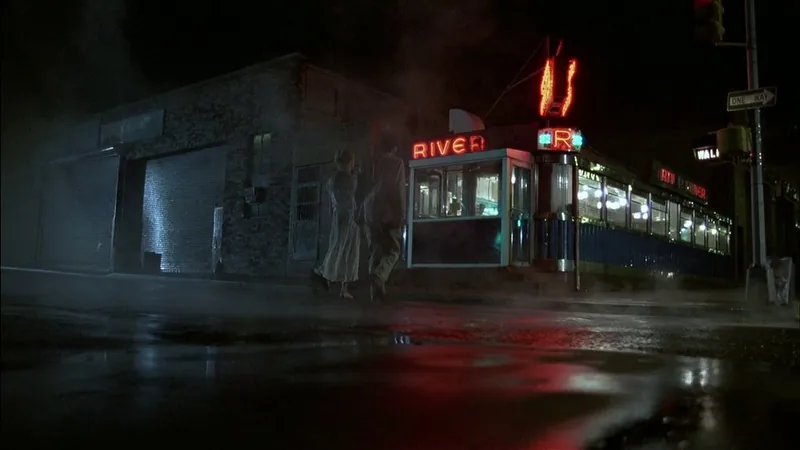 Two figures walk past a diner lit with red neon on a misty wet city street.