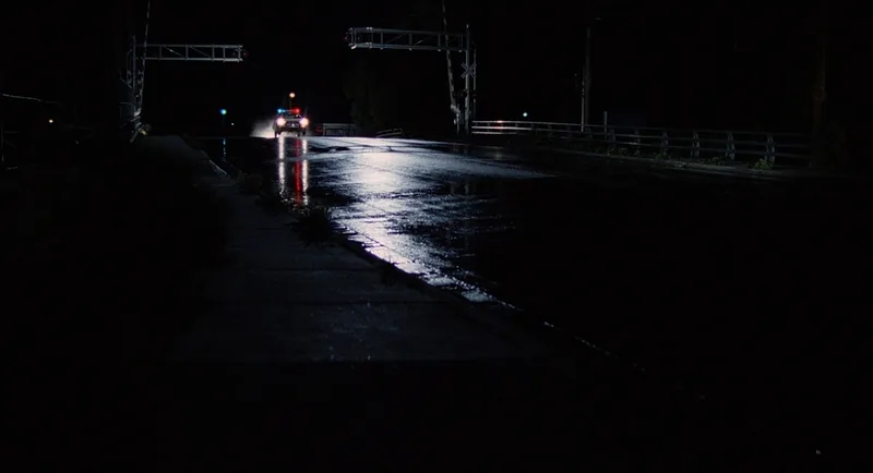 Car headlights cast long white reflections along a rain-slicked empty road under overhead gantry trusses.